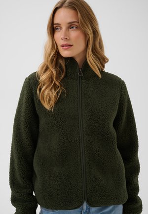 Femme aux longs cheveux blonds portant une veste polaire zippée texturée vert foncé et un jean bleu clair, regardant légèrement sur le côté.