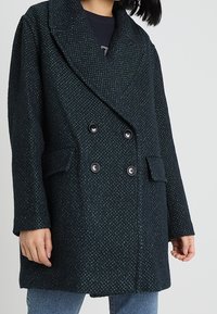 Abrigo de doble botonadura en verde oscuro, fabricado con un tejido texturizado, con grandes solapas, dos bolsillos frontales y detalles de botones negros.