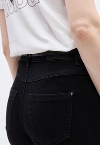 Κοντινό πλάνο ενός ατόμου που φοράει μαύρο τζιν και λευκό πουκάμισο, εμφανίζοντας λεπτομέρειες της πίσω τσέπης και μέρος του χεριού με ορατή υφή δέρματος στον αγκώνα.