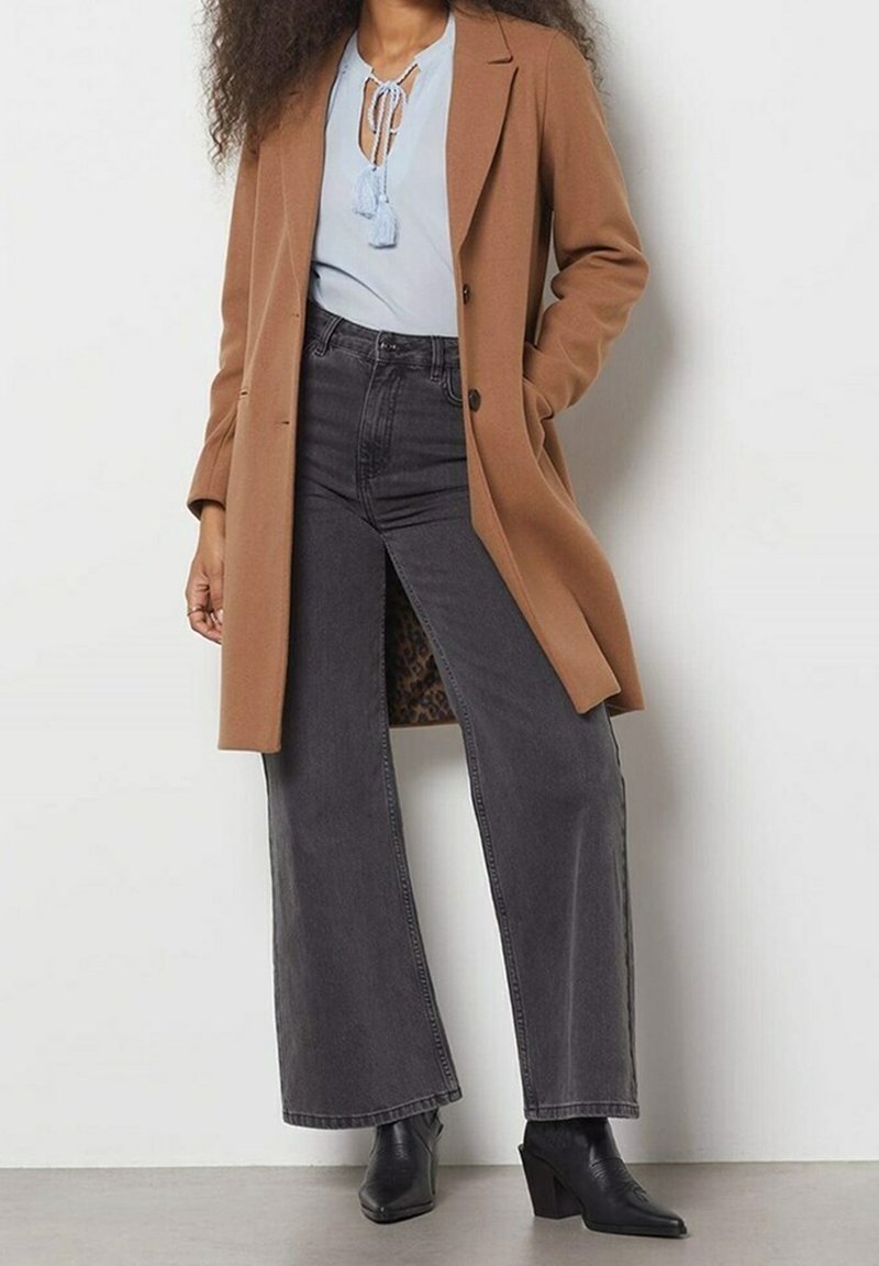 Femme portant un long manteau marron, un chemisier bleu clair à franges, un jean noir taille haute à jambes larges et des bottes noires à talons, debout contre un mur blanc.