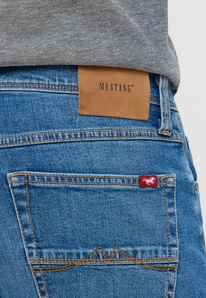Mėlynos džinsinės kelnės su ruduo odiniu etiketę, ant kurios užrašyta "MUSTANG", ir raudona etikete su arklių logotipu. Išskirtinės siuvimo detalės ant kišenių.