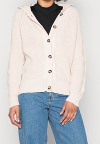 Jasno beżowy sweter dziany z kapturem, z ribbowanymi mankietami i zapięciem na guziki z przodu z drewnianymi guzikami. Widoczna textura w geometryczny wzór.