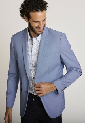 Homme souriant ajustant un blazer bleu clair sur une chemise blanche à motifs et un pantalon foncé sur un fond uni.