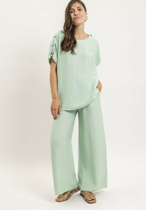 Femme portant un pantalon ample vert clair et un haut à manches courtes assorti avec des détails de boutons sur les épaules, debout avec une main dans la poche.