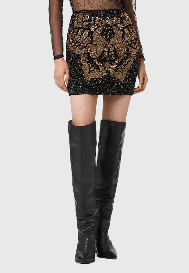 Czarna mini spódnica z cekinami i złotymi ozdobami oraz skomplikowanymi wzorami, w zestawie z dopasowanymi czarnymi kozakami do kolan.