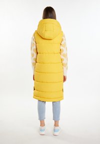 Gilet imbottito giallo con cappuccio, caratterizzato da cuciture orizzontali e tasche laterali, abbinato a jeans azzurri chiari e sneakers multicolori.