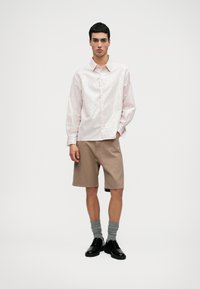 Svetloružová košeľa na gombíky s vertikálnymi prúžkami, skombinovaná s béžovými šortkami. Model má na sebe šedé ponožky a čierne topánky, stojí na jednolatom pozadí.