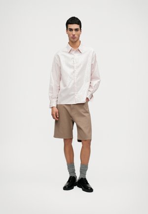 Gaišs rozā krekls ar vertikālām svītrām, apvienots ar bēšiem šortiem. Modelis valkā pelēkus zeķes un melnas kurpes, stāv uz neitrālā fona.