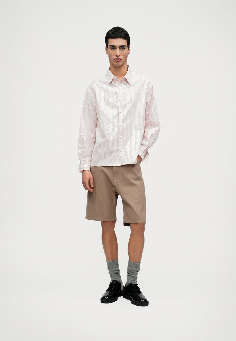 Svetloružová košeľa na gombíky s vertikálnymi prúžkami, skombinovaná s béžovými šortkami. Model má na sebe šedé ponožky a čierne topánky, stojí na jednolatom pozadí.