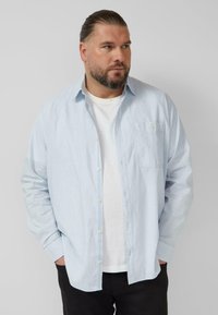 Hellblaues Hemd mit Knöpfen und Brusttasche, darüber ein weißes T-Shirt getragen. Eng anliegende Passform, lange Ärmel, glatte Baumwollstruktur.