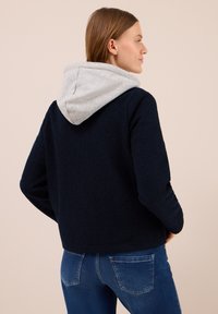 Nainen, jolla on vaaleanruskeat hiukset, pukeutuneena mustaan fleece-takkiin, jossa on harmaa huppu, yhdistettynä sinisiin farkkuihin, seisoo yksinkertaista taustaa vasten.