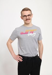 Šedé bavlněné tričko s multibarevným grafikou "JT Racing". Nosené s černými kalhotami a černým opaskem. Hladká textura, krátké rukávy.