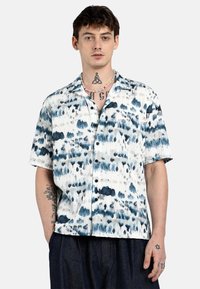Camicia a maniche corte con bottoni di colore bianco, decorata con motivi tie-dye blu e grigi. Presenta una vestibilità ampia e un colletto, con una finitura in tessuto testurizzato.