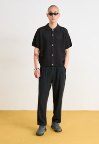 Black knit polo shirt with short sleeves and buttons, paired with loose black trousers and gray sneakers. Smooth texture, minimal design.