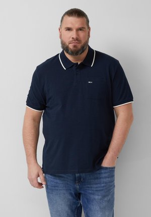 Brodaty mężczyzna w granatowej koszulce polo z białym obszyciem i niebieskich dżinsach, stojący z jedną ręką w kieszeni na jednolitym tle.