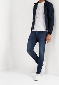 Chaqueta ligera de marina sobre una camiseta blanca con un pequeño estampado, combinada con jeans azul oscuro y zapatillas blancas. Un atuendo casual con un diseño sencillo.