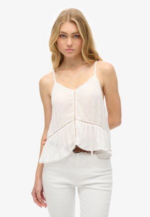 Femme blonde portant un haut blanc sans manches avec des détails en dentelle et un pantalon blanc, posant devant un fond clair uni.