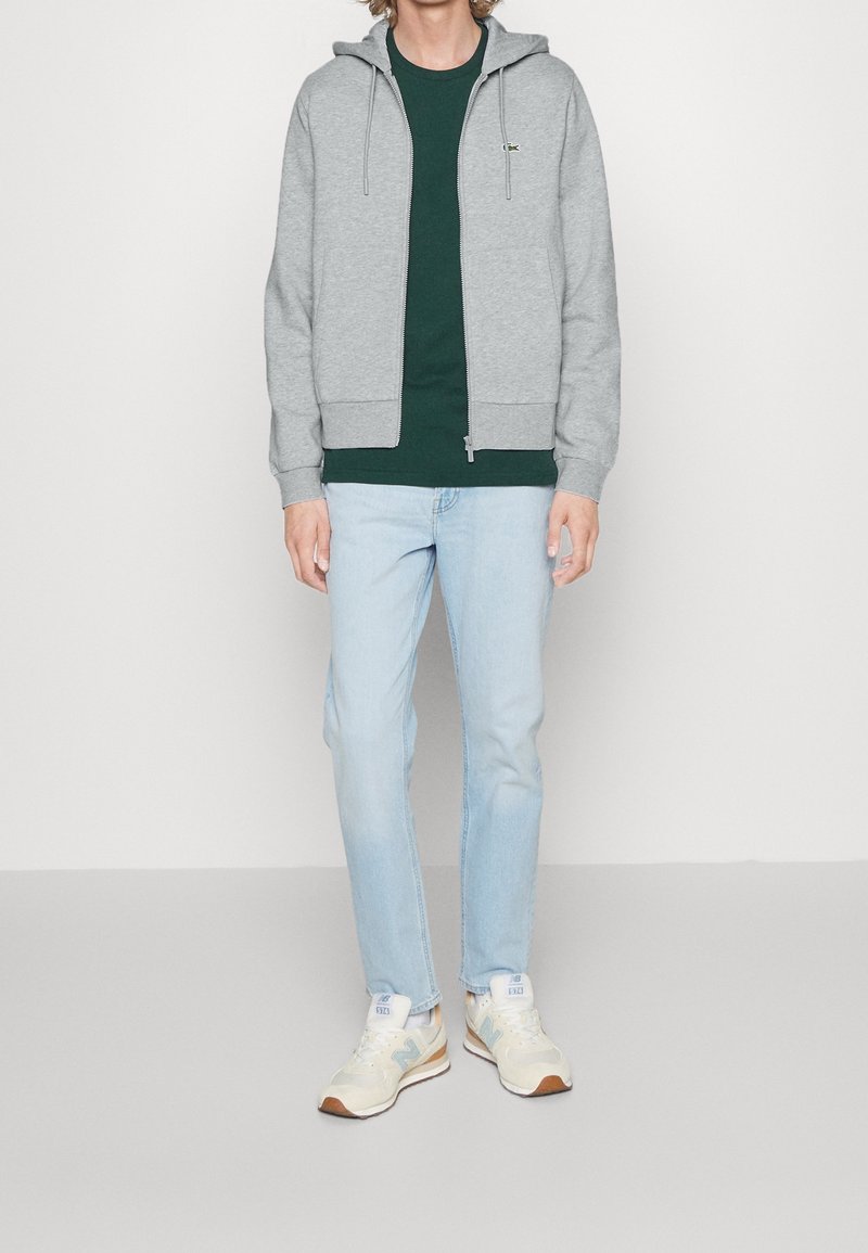 Personne portant un sweat à capuche gris zippé, un t-shirt vert foncé, un jean bleu clair et des baskets beiges, debout devant un fond clair uni.
