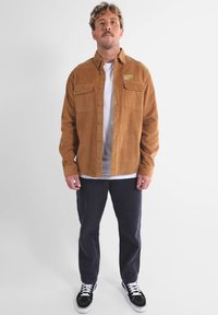 Veste chemise en velours côtelé marron avec deux poches poitrine, boutonnière à l'avant, portée sur un t-shirt blanc, associée à un pantalon noir et des baskets.