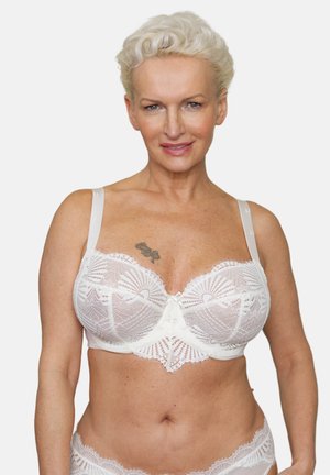 Mature woman with short white hair wearing a white lace bra and matching underwear, standing against a plain background.