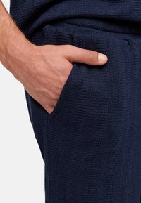 Marineblaue Struktur-Hosen mit einer Hand in der Tasche. Das Material wirkt weich und gewebt, mit sichtbaren Nähten entlang der Kanten.