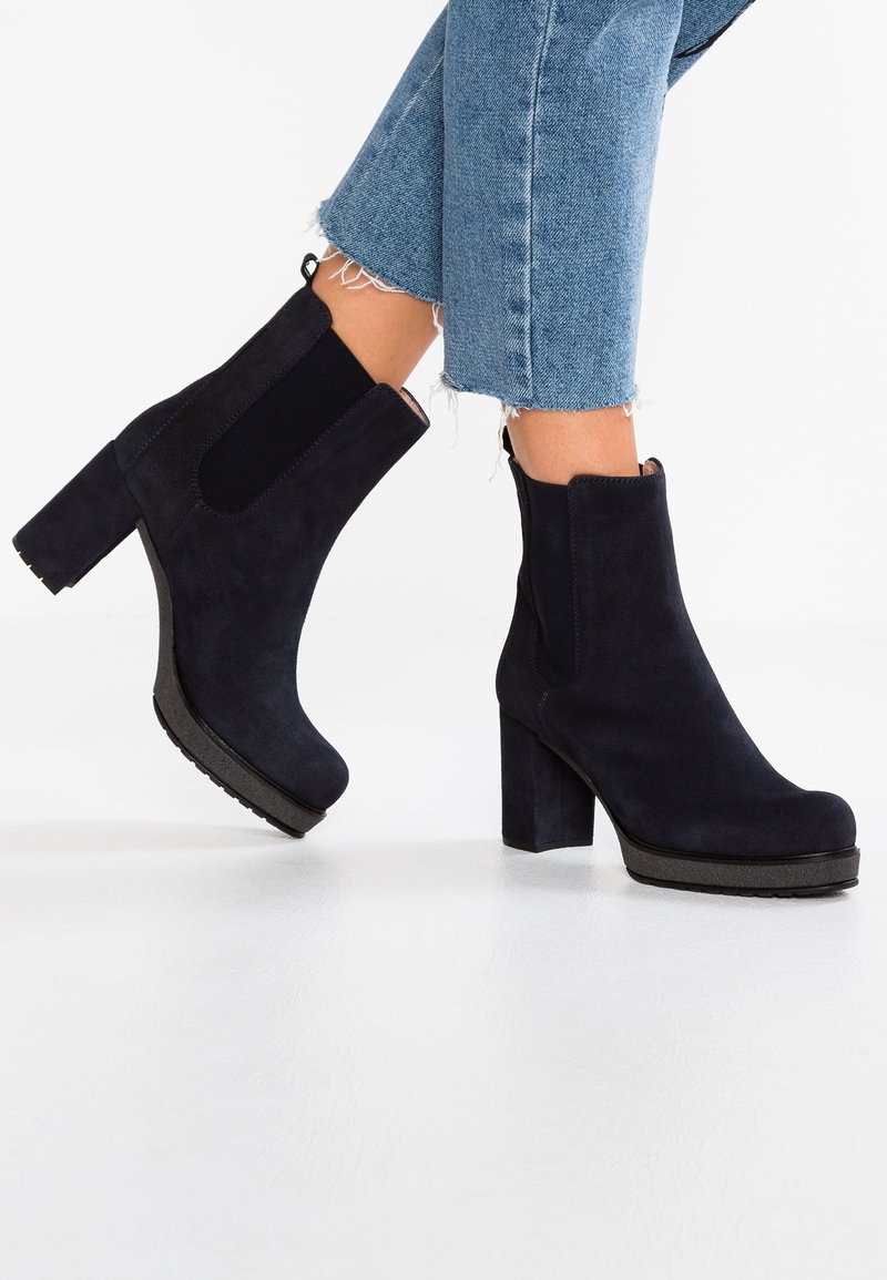 Des pieds chaussés de bottines à talons blocs en suède noir, associées à un jean léger bleu clair avec des ourlets effilochés.