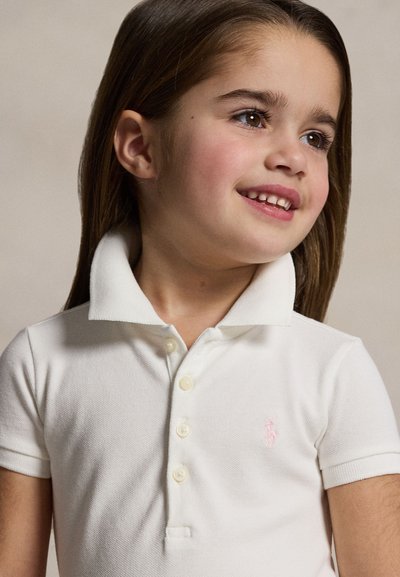 Young girl with long brown hair smiles while wearing a white collared short-sleeve shirt against a plain background.