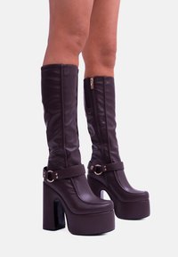 Stivali alti fino al ginocchio in finta pelle marrone con platform e tacco spesso, dotati di una zip laterale e di una cinghia regolabile con dettagli in metallo.