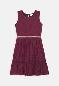 Mouwloze bourgondische jurk met kleine witte stippen, een geplooide rok met ruches, en een dunne gouden riem om de taille.