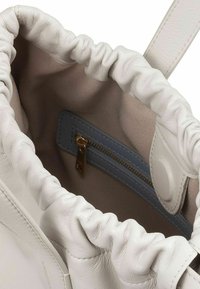 Borsa in pelle bianca con parte superiore arricciata, che mostra una fodera beige e una tasca interna blu con zip dorata. Texture soffice, design funzionale.