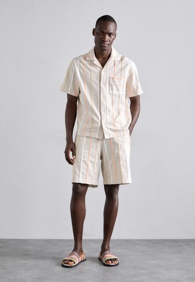 Helles beiges kurzärmliges Hemd und passende Shorts mit orangefarbenen und gelben vertikalen Streifen. Getragen mit mehrfarbigen gestreiften Sandalen.