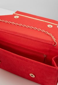 Červená látková kabelka se zlatým kováním, řetízkovým popruhem a vnitřní kapsou na zip. Hladká textura s knoflíkovým zapínáním.