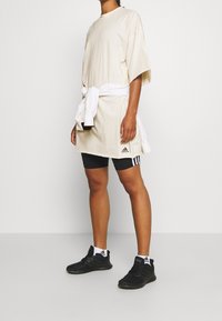 Camiseta de gran tamaño beige con mangas cortas, con una tela blanca atada en la cintura, combinada con pantalones cortos negros y zapatillas deportivas negras.