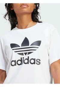 Camiseta de algodón blanca que presenta un gran logo negro de Adidas con tres rayas y un diseño de hoja. Mangas cortas y escote redondo.