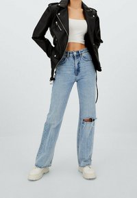 Czarna skórzana kurtka, biała crop top, dżinsy w średnim kolorze, wysoki stan z przetarciami oraz masywne, białe sneakersy w odcieniu ecru.