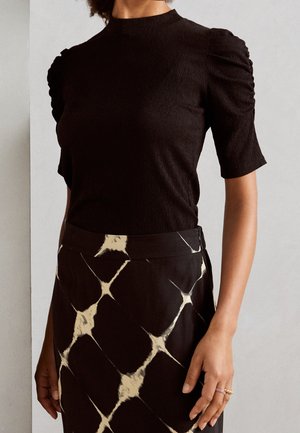 Kvinde iført en sort tekstureret kortærmet bluse med pufærmer og en sort nederdel med beige abstrakt tie-dye mønster, hånd synlig med ringe.