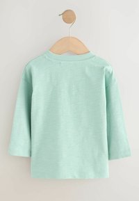 Chemise à manches longues vert clair suspendue sur un cintre en bois. Le tissu présente une texture subtile avec un col rond et un ourlet droit.