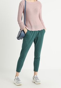 Ljusrosa stickad tröja med ribbade muddar och vågformad hem, kombinerad med gröna tapered byxor. Accessoarer med en blå axelväska och vita sneakers.