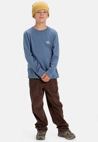 Modrá košeľa s dlhými rukávmi s motívom hory, doplnená hnedými manšestrovými nohavicami a žltou rebrovanou čiapkou; neformálne atletické topánky.