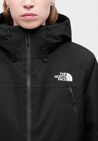 Černá bunda s vysokým límcem a nastavovací kapucí. Obsahuje zipový přední uzávěr a boční kapsy. Logo na hrudi v bílé barvě. Hladká textura.