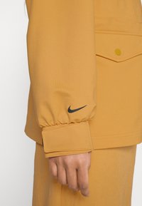Gul Nike-jacka med elastisk mudd, med en svart logotyp. Slätt tyg och enkel design med en detalj av en knappficka.