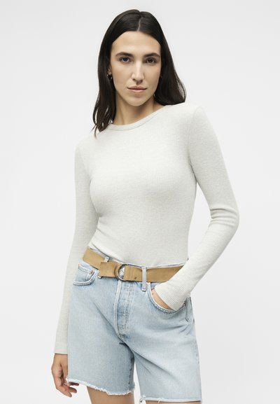 Νεαρή γυναίκα με εφαρμοστή, ανοιχτό γκρι μπλούζα με μακριά μανίκια, ανοιχτό μπλε τζιν σορτς με ξεφτισμένα τελειώματα και μπεζ ζώνη, ποζάρει με το ένα χέρι στην τσέπη.