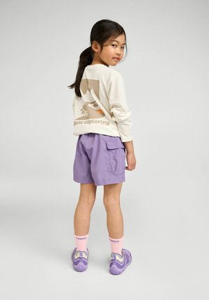 Dziewczyna z ciemnymi włosami związanymi w kucyk, ubrana w białą koszulę z długim rękawem z napisem "gone exploring", fioletowe szorty, różowe skarpetki i fioletowe tenisówki.