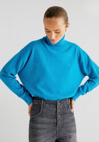 Hellblauer Rollkragenpullover aus weichem Material, mit lässiger Passform und gerippten Bündchen, kombiniert mit hochtaillierten, dunkelgrauen Jeans.