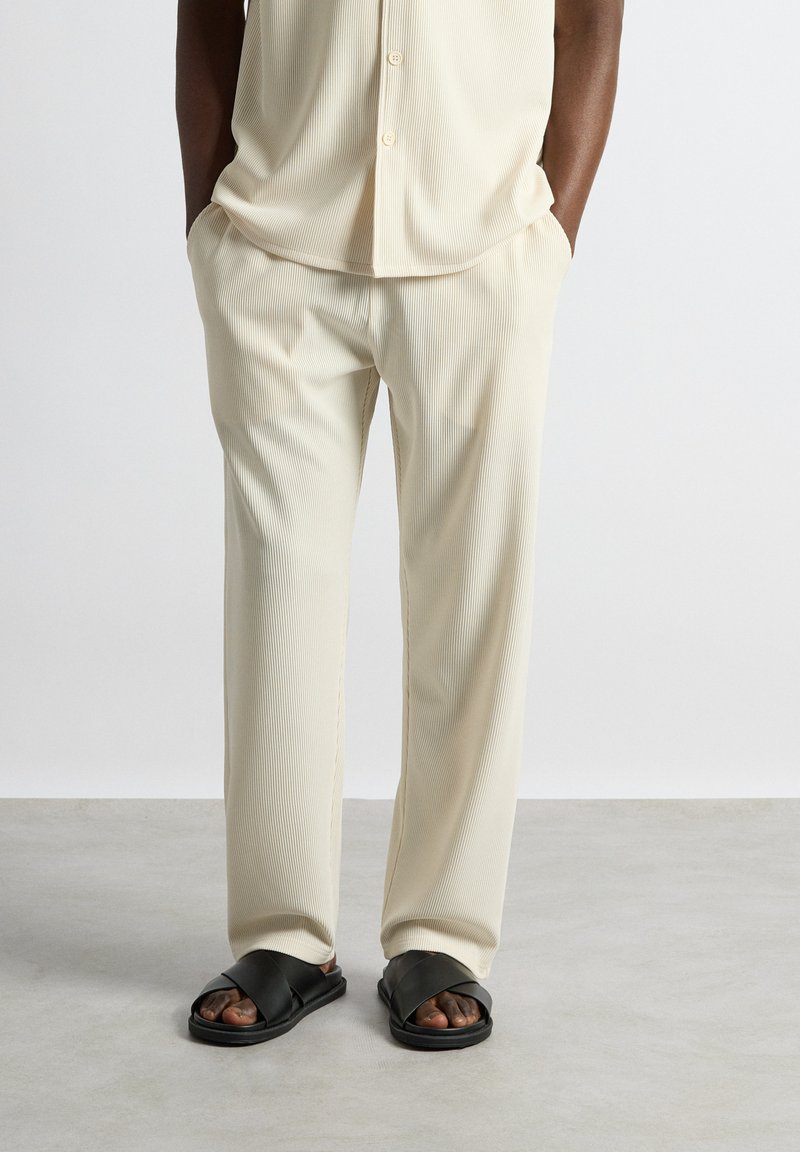 Homme portant une chemise crème côtelée à boutons et un pantalon assorti avec des sandales noires croisées, mains dans les poches, debout sur un sol gris.