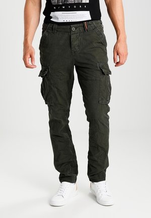 Männliches Model trägt dunkelgrüne Cargo-Hosen und weiße Sneakers, steht vor einem einfarbigen hellen Hintergrund.