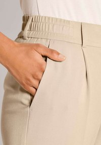 Hand rustend in de voorzak van beige high-waisted broek met een elastische tailleband en riemlussen.