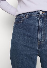 Mörkblå denimjeans med hög midja, med framfickor och kontrasterande sömmar. Slät struktur och klassisk passform.