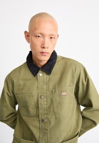 Grøn bomuldsjakke med en sort corduroy-krave, frontknapper, to brystlommer og et logo-patch på den venstre lomme.