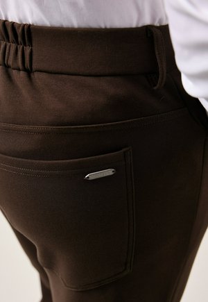 Nahaufnahme von dunkelbraunen Hosen mit elastischem Bund und Gesäßtasche, die ein kleines Metalletikett mit dem Text "JP 1880" trägt.